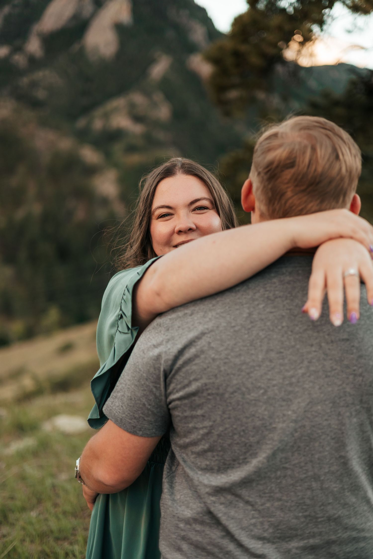 A smiling woman in a green dress hugs a man outdoors with mountain scenery  | Baby Announcement Photographer in Boulder, Colorado