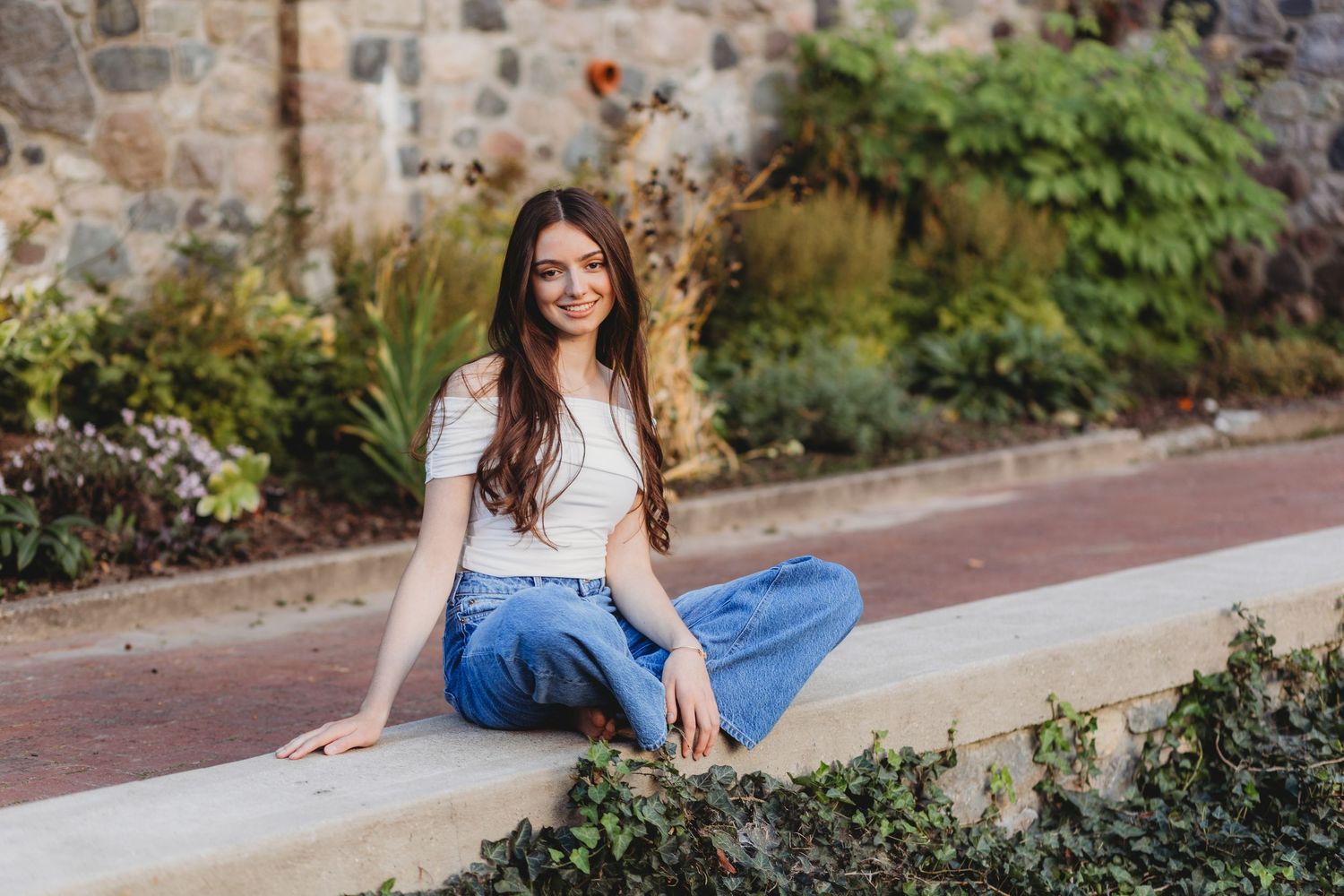 Young woman with long brown hair smiling, seated on a stone ledge in a garden wearing a white top and blue jeans. cranbrook senior session