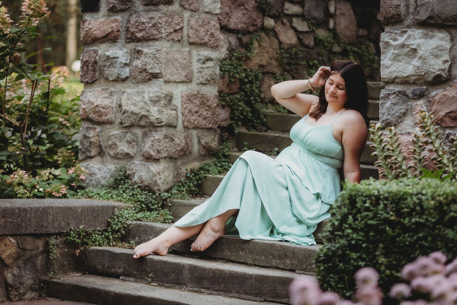A woman in a mint green dress poses gracefully on stone steps surrounded by lush garden greenery. Birmingham senior session