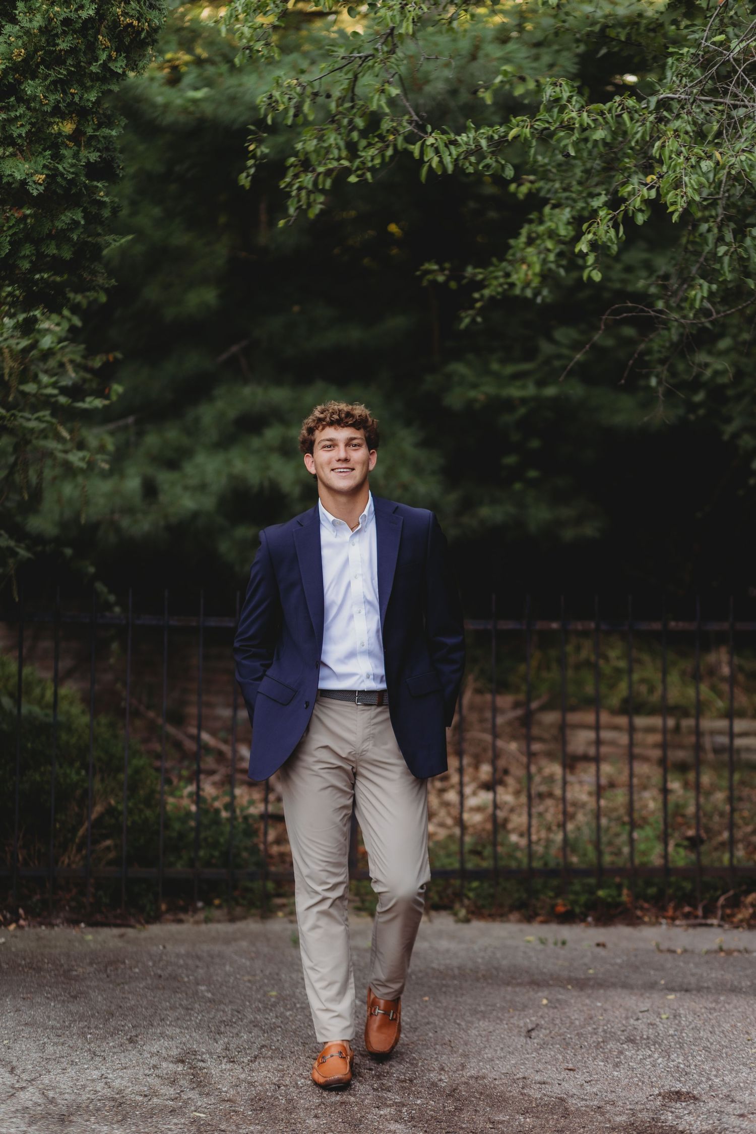 Young man in navy blazer and khaki pants smiling outdoors near green trees and iron fence. regina high school st Hugo church