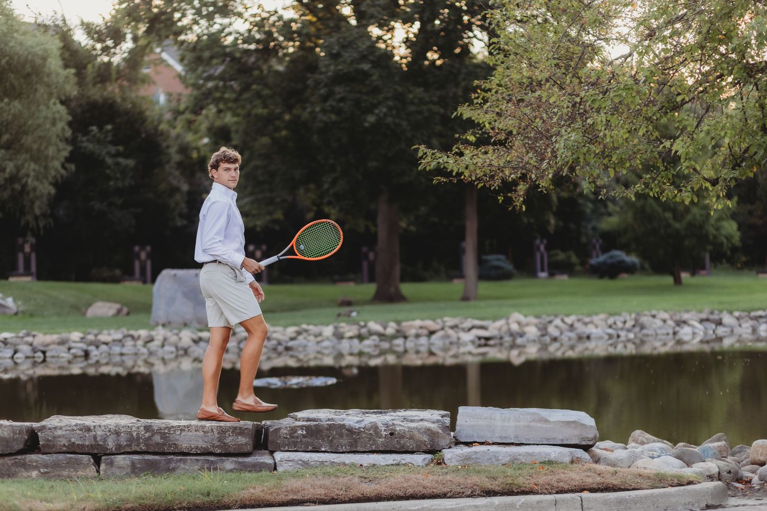 A young boy holding a tennis racket stands on rocks beside a serene pond in a lush green park. senior session st Hugo church