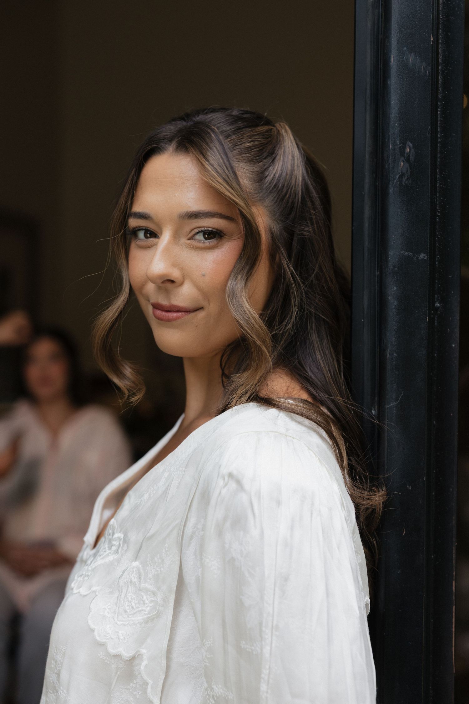 Young woman in white embroidered blouse leaning against dark doorframe, smiling softly with wavy brown hair.