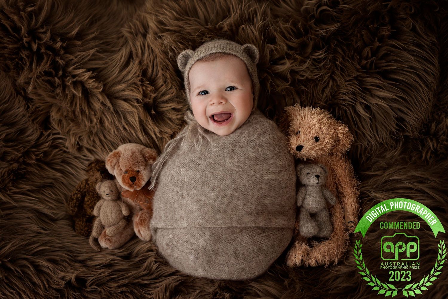 Smiling baby wrapped in brown bear bonnet surrounded by teddy bears on fluffy fur, APP award-winning newborn photo 2023.
