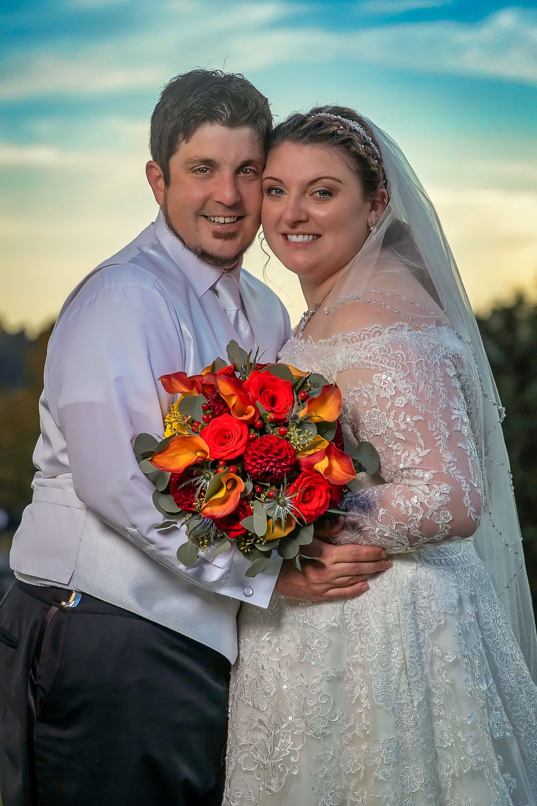 Bride and groom smiling together outdoors, bride holding vibrant red and orange rose bouquet, wearing lace gown and veil.