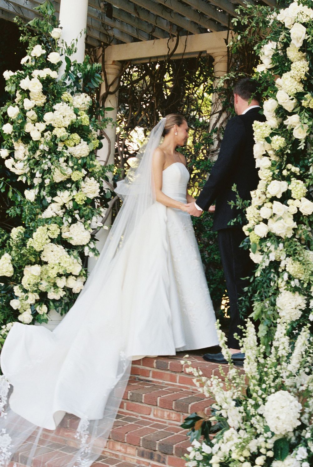 Bride and groom walk through stunning white floral arch at outdoor wedding ceremony.