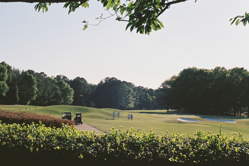 Lush golf course fairway with sand bunkers, golf carts, and players surrounded by tall trees on a clear sunny day.