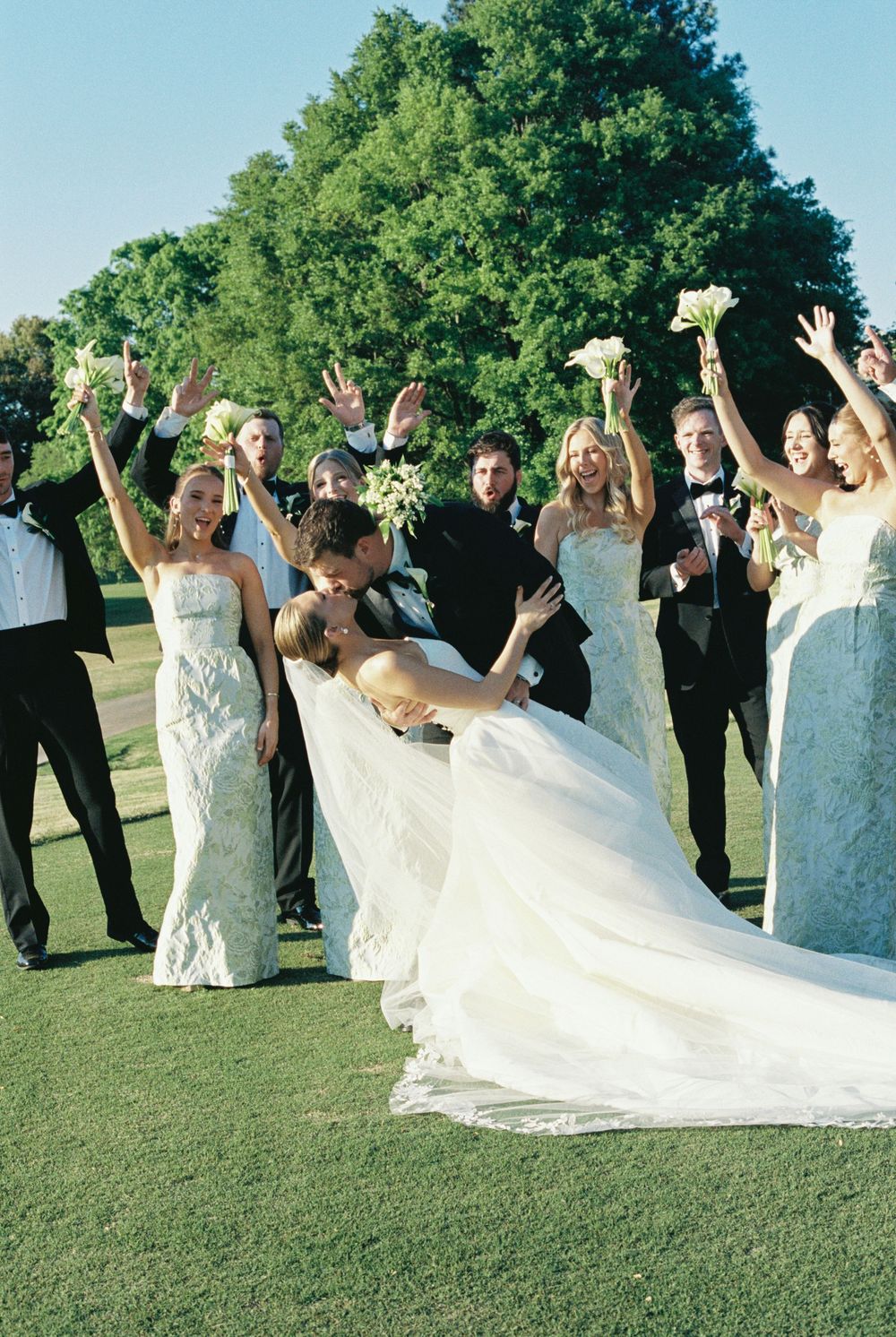 Joyful wedding party cheers as groom dips bride for a kiss on lush green golf course on sunny day.
