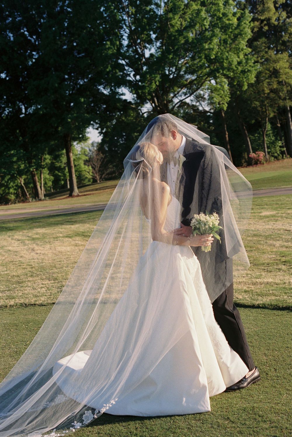 Bride and groom share romantic kiss under flowing veil on lush green lawn.