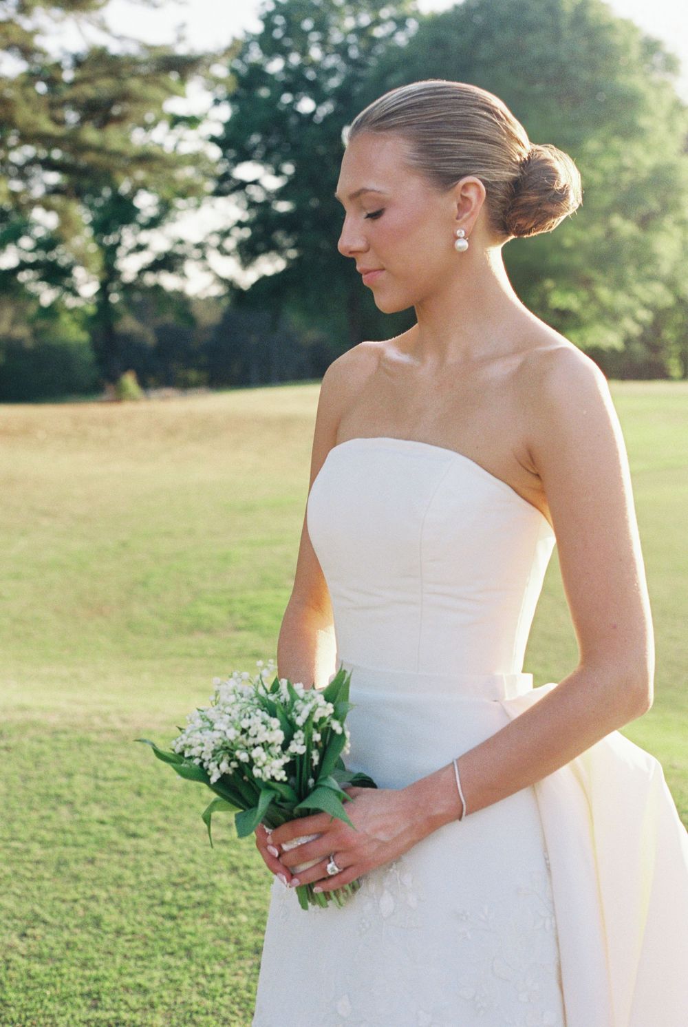 Elegant bride in a strapless gown holding a small lily of the valley bouquet in golden outdoor light.