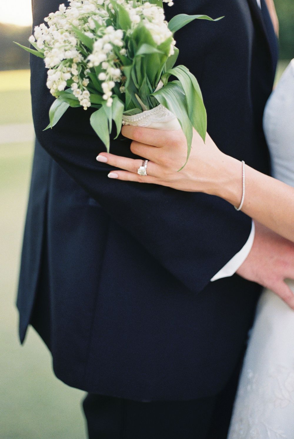 Bride holding lily of the valley bouquet with diamond ring visible against grooms dark navy suit.