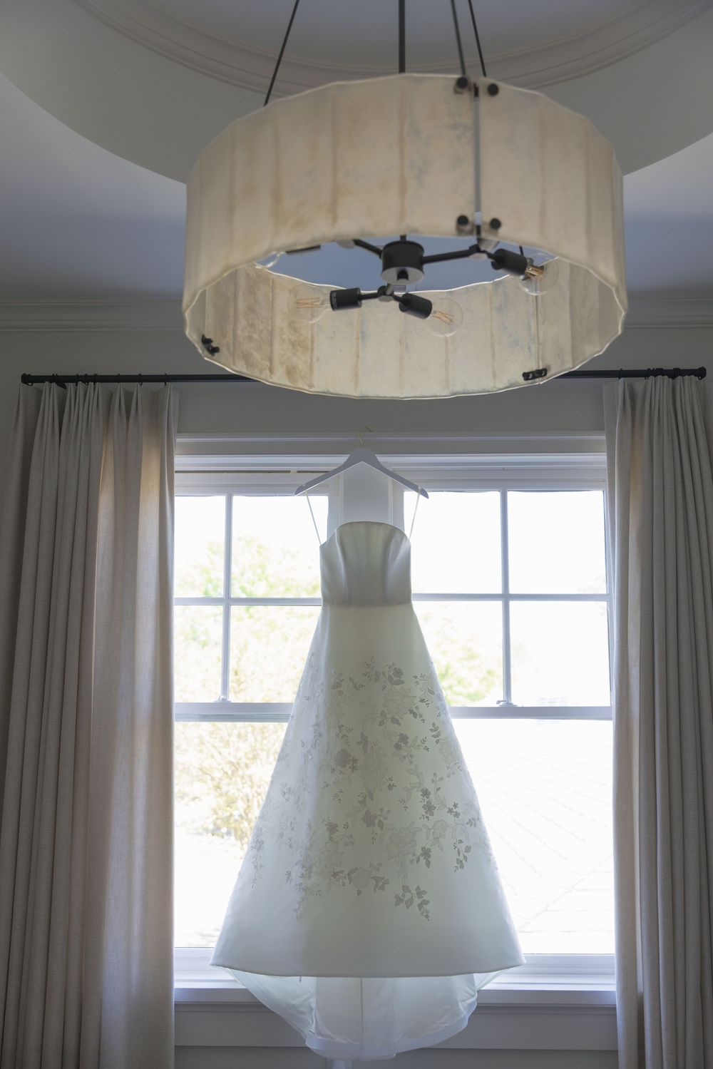 Elegant white floral wedding gown hanging in front of a sunlit window beneath a drum chandelier.