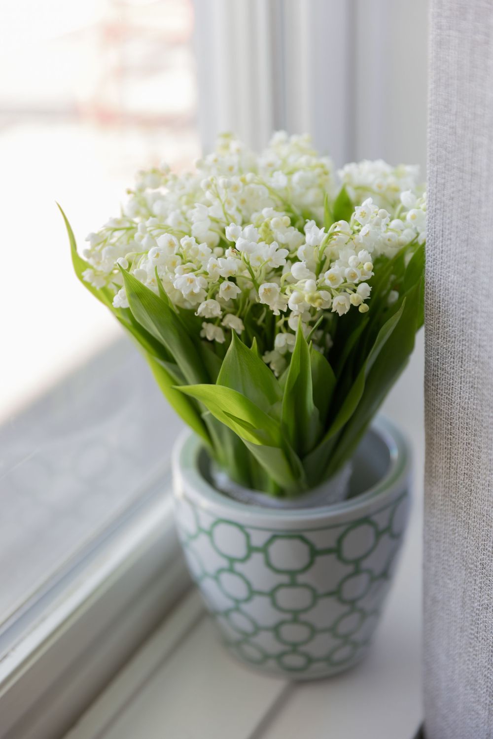 Fresh lily of the valley bouquet in a green geometric patterned vase on a white windowsill.