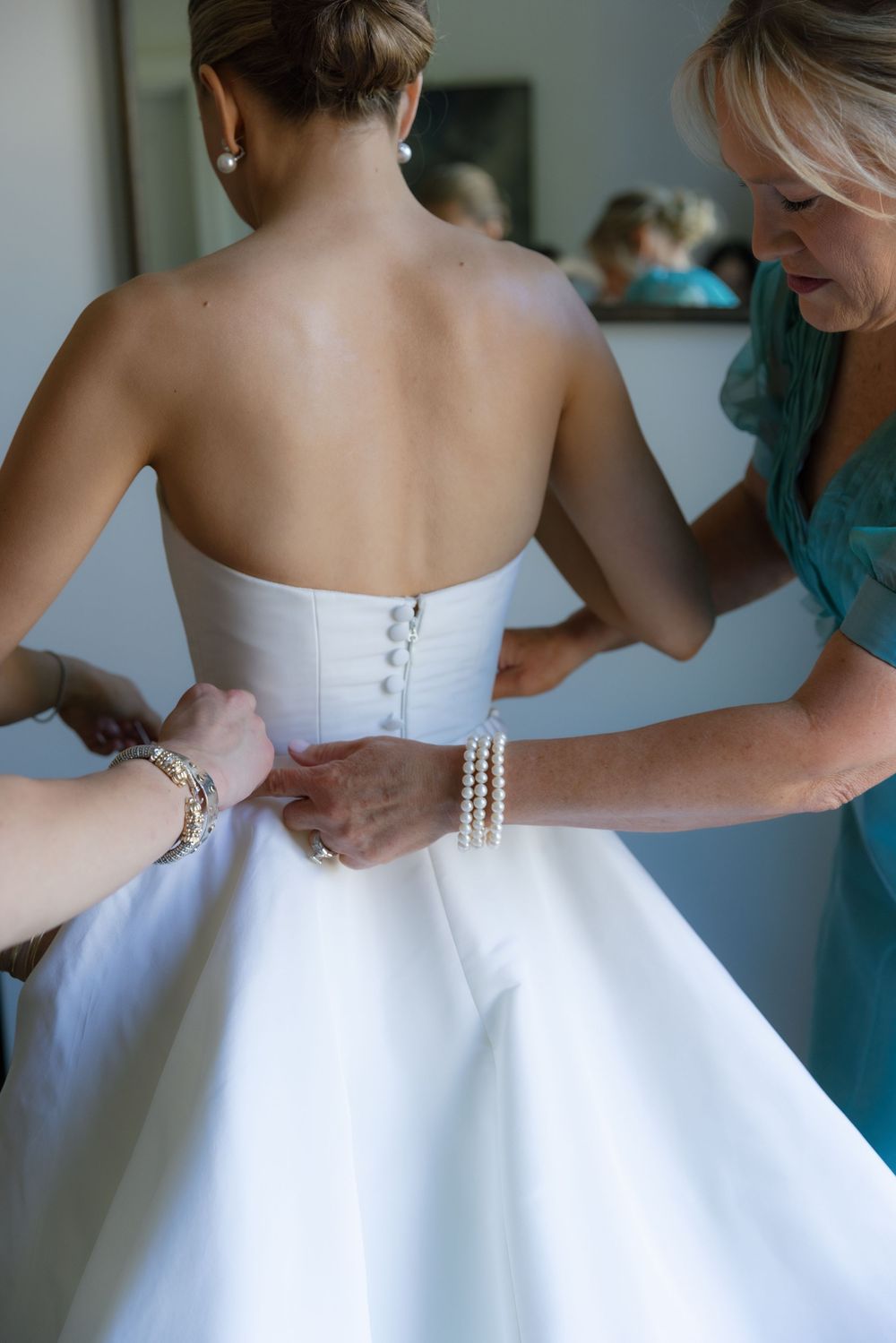 Bride being helped into a strapless white wedding dress with button back by bridesmaids.