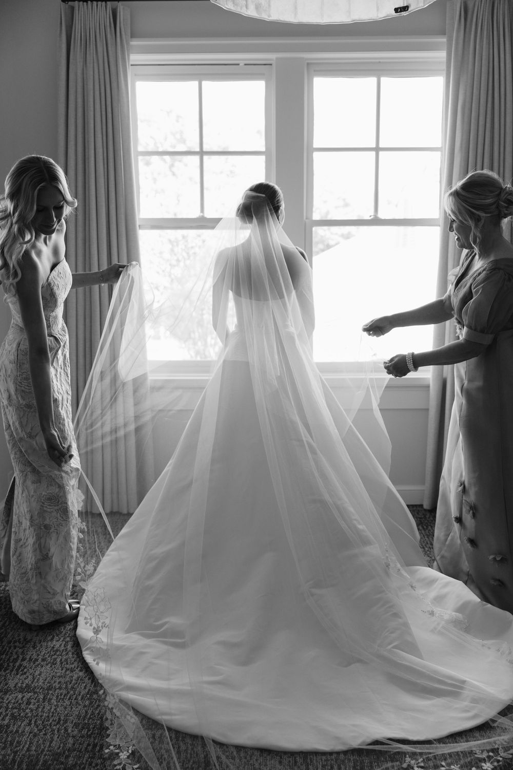 Bride in elegant white gown and veil stands by window as two bridesmaids arrange her train in black and white photo.