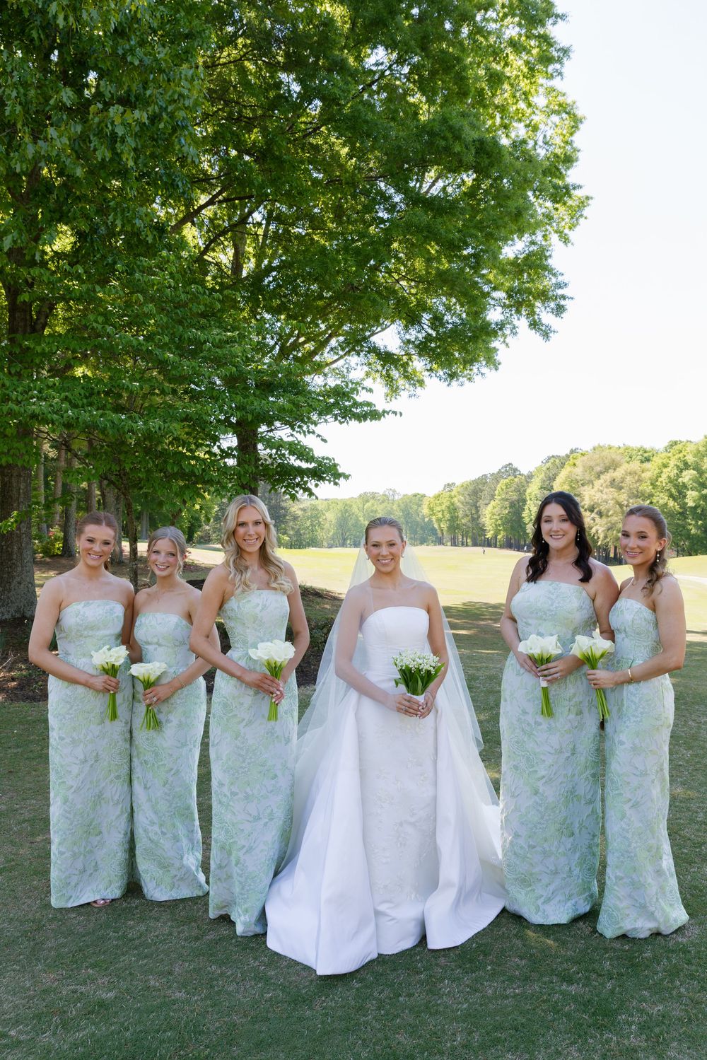 Bride in white gown poses with five bridesmaids in sage green lace dresses outdoors.