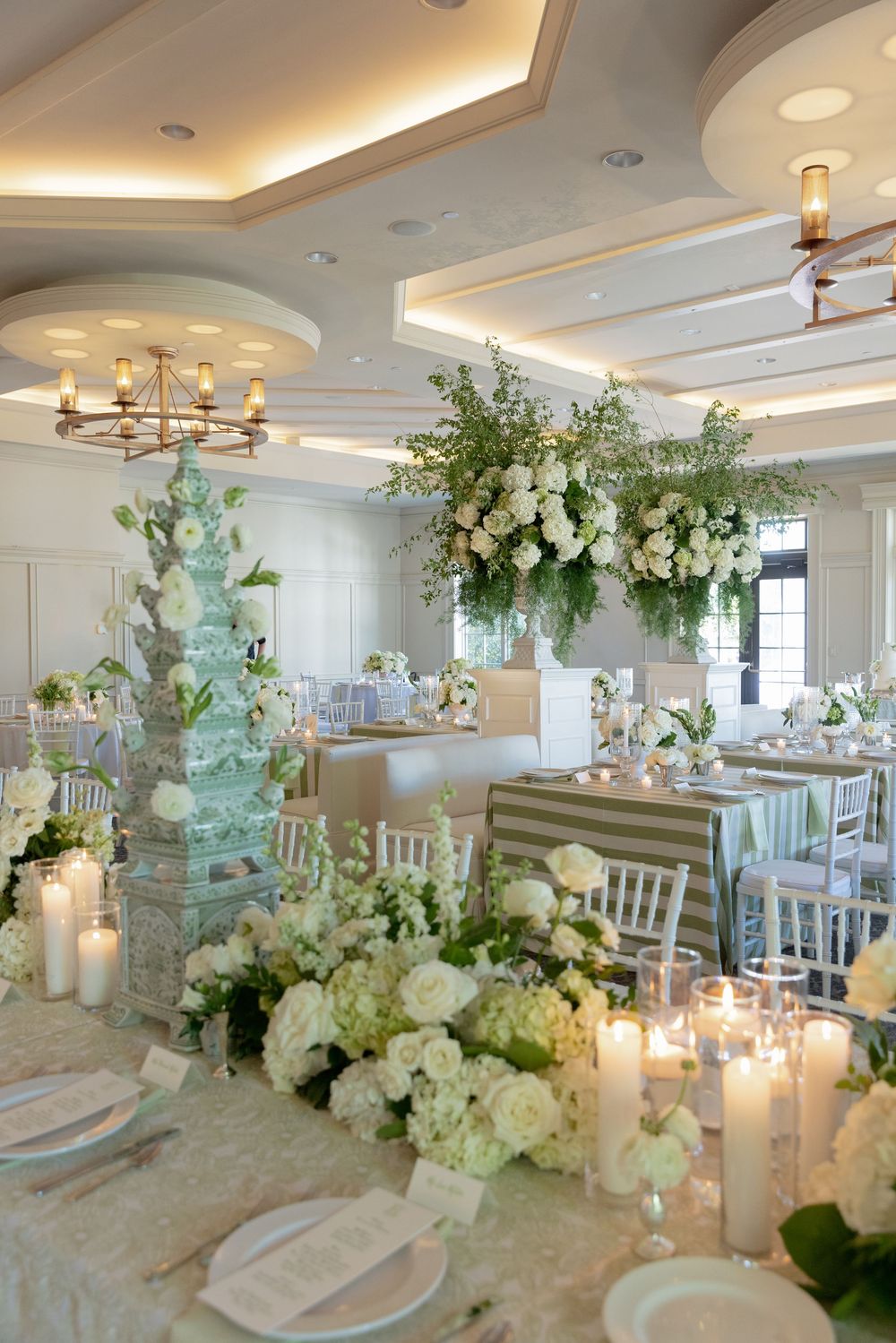 Elegant wedding reception hall with white floral arrangements, candles, and a tiered cake in a softly lit ballroom.