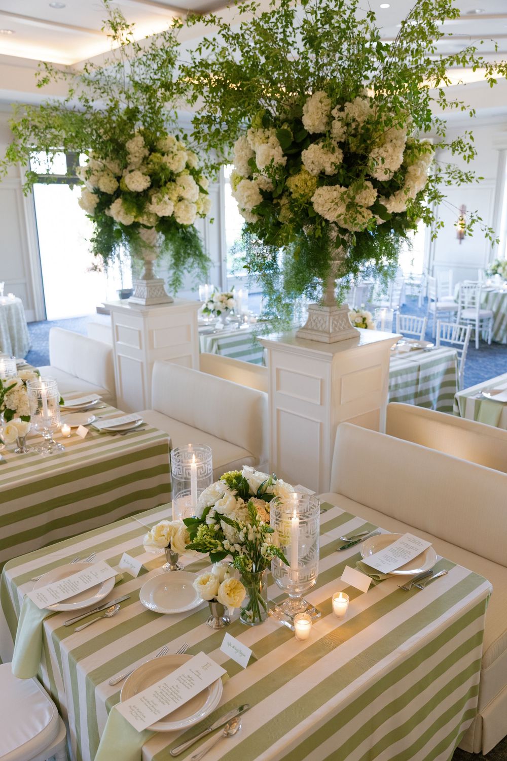 Elegant wedding reception tables with green striped linens, white floral centerpieces, candles, and lush greenery arrangements overhead.