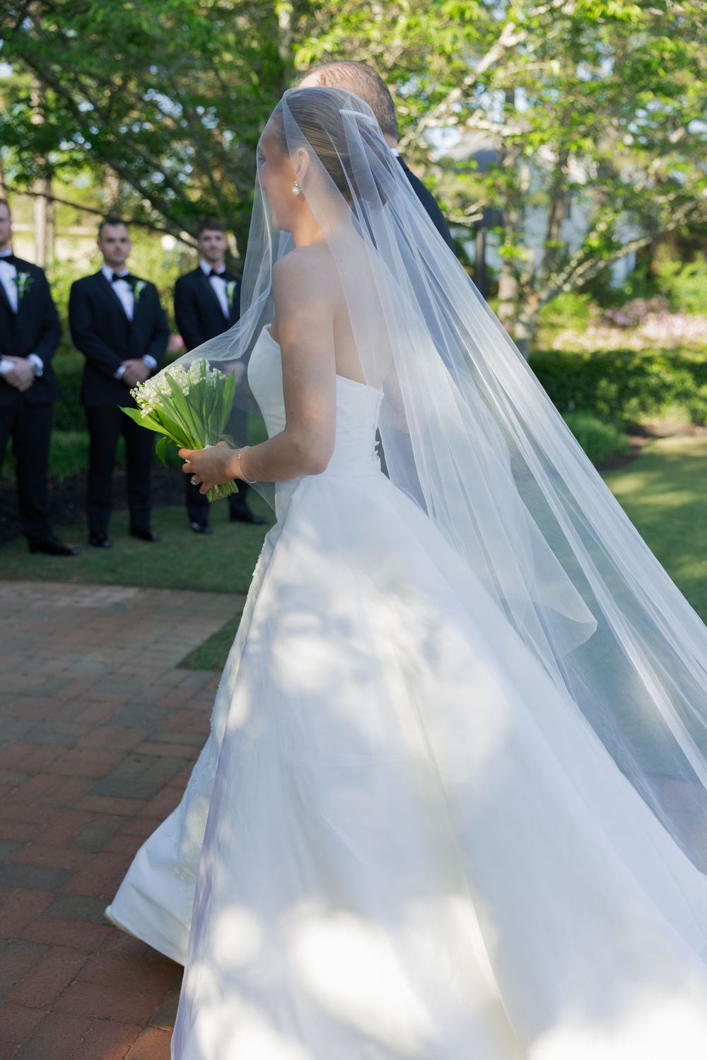 Bride in white strapless gown and veil holding bouquet, with groomsmen in black tuxedos standing in background.