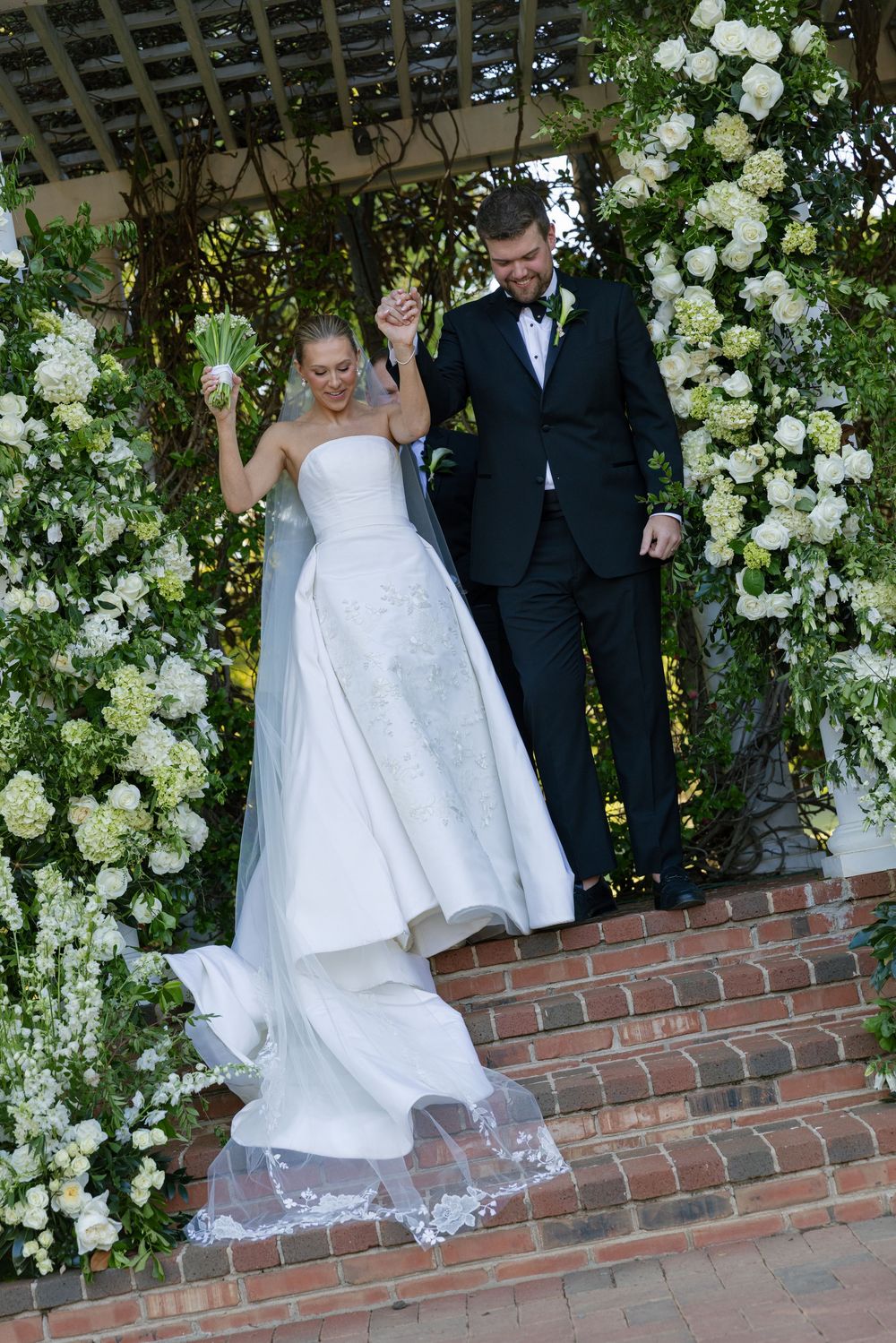 Bride and groom celebrate on brick steps beneath a lush floral arch of white hydrangeas and greenery.