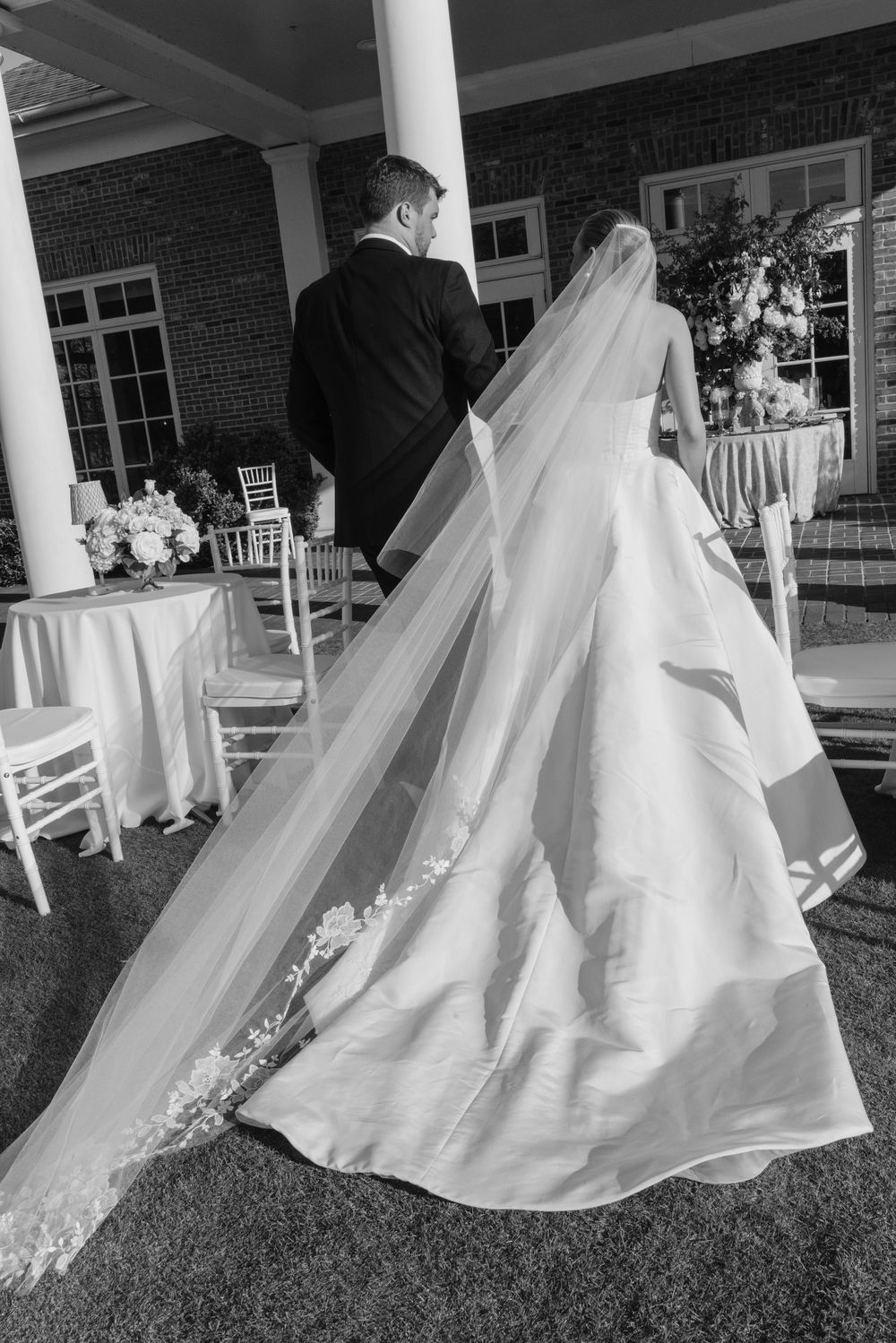 Black and white photo of bride and groom walking toward elegant outdoor wedding reception venue with columns.