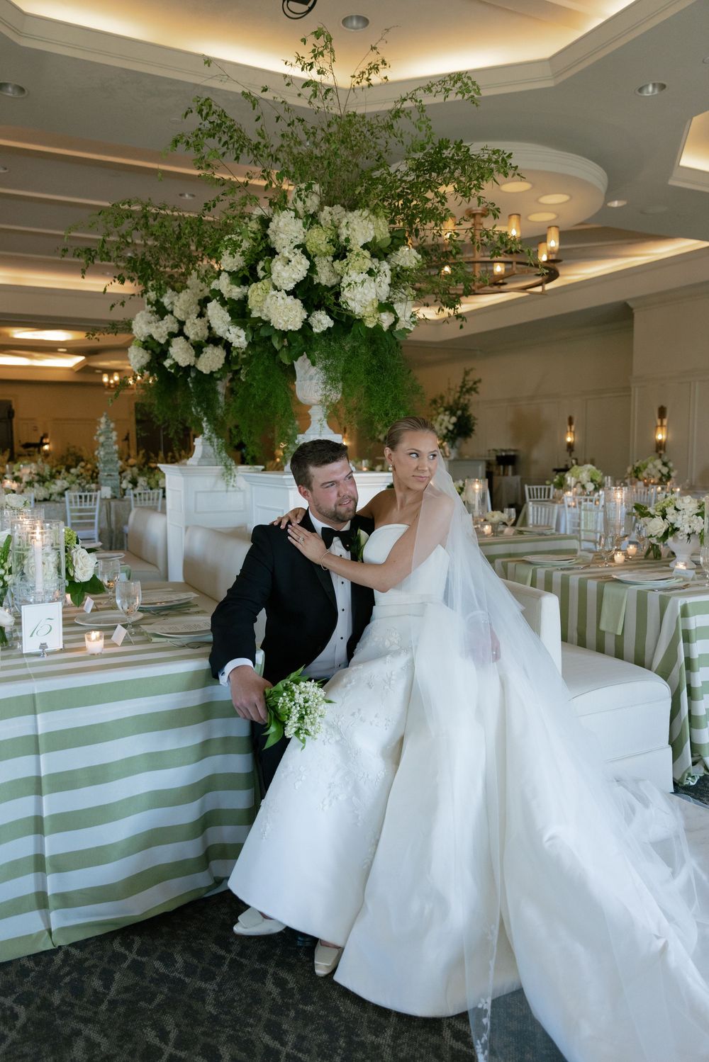 Bride and groom pose at sweetheart table adorned with green striped linens and lush floral centerpiece.