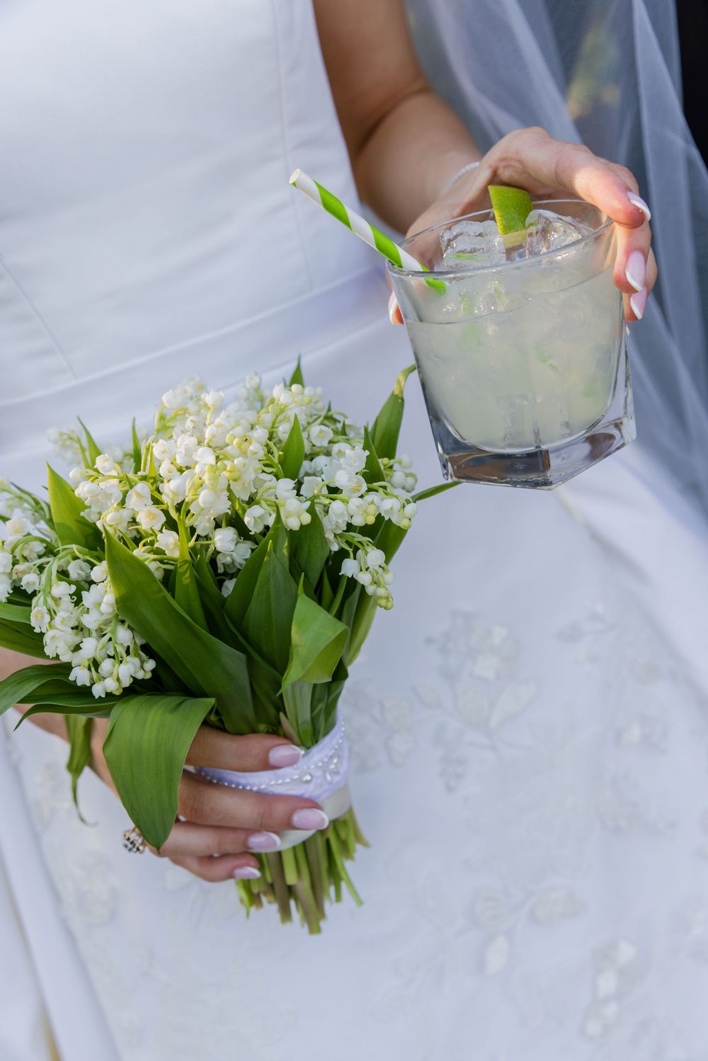 Bride holding a lily of the valley bouquet and an iced cocktail with a green straw in her hands.