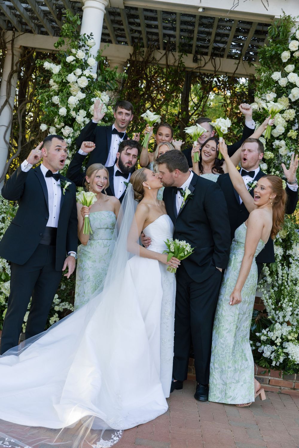 Joyful wedding party celebrates with bride and groom kissing under flower-covered gazebo in outdoor ceremony.