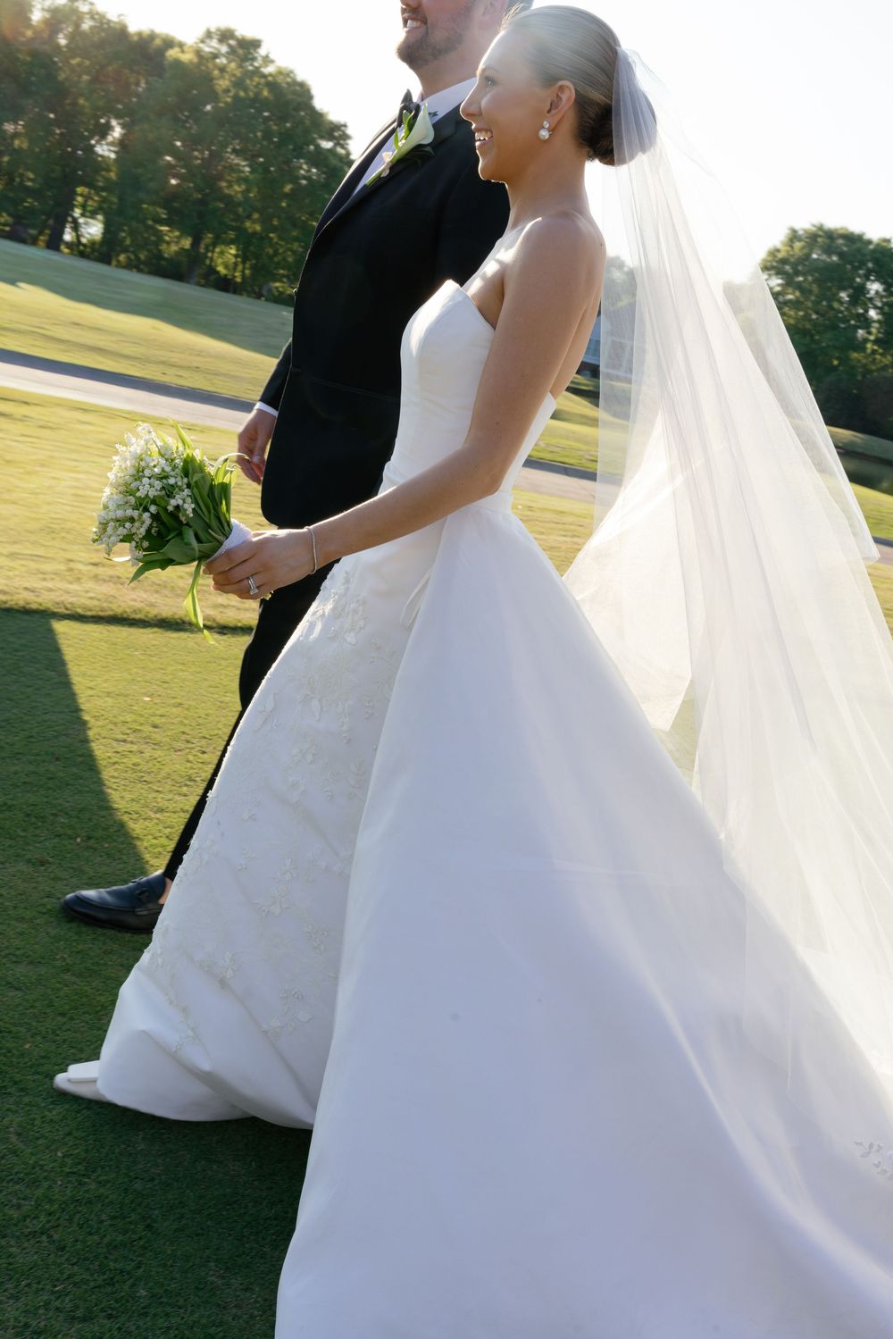 Bride in a flowing white strapless gown and veil walks hand-in-hand with groom in black suit on a green lawn.