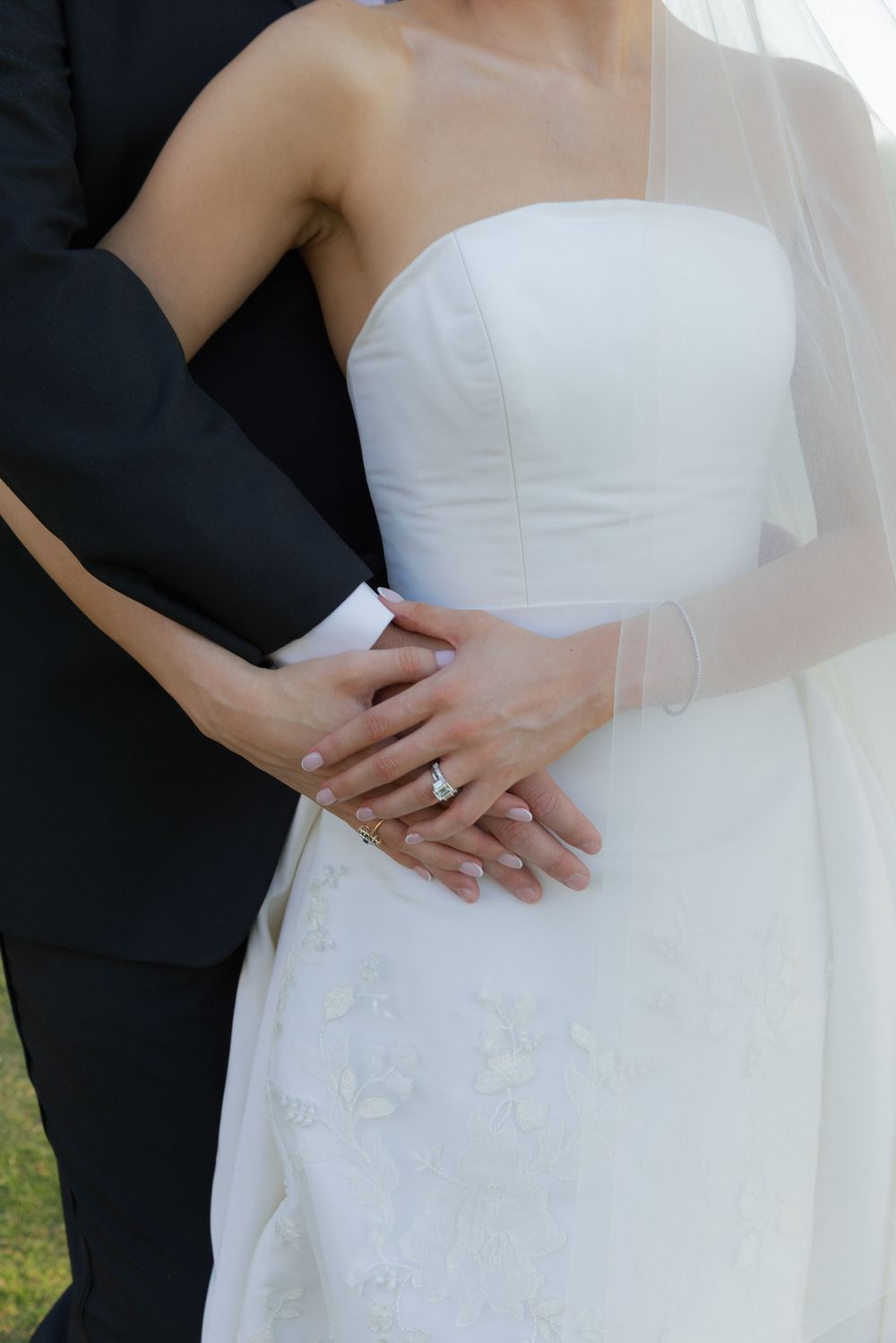 Close-up of bride and groom hands showing diamond engagement ring against white strapless wedding gown.