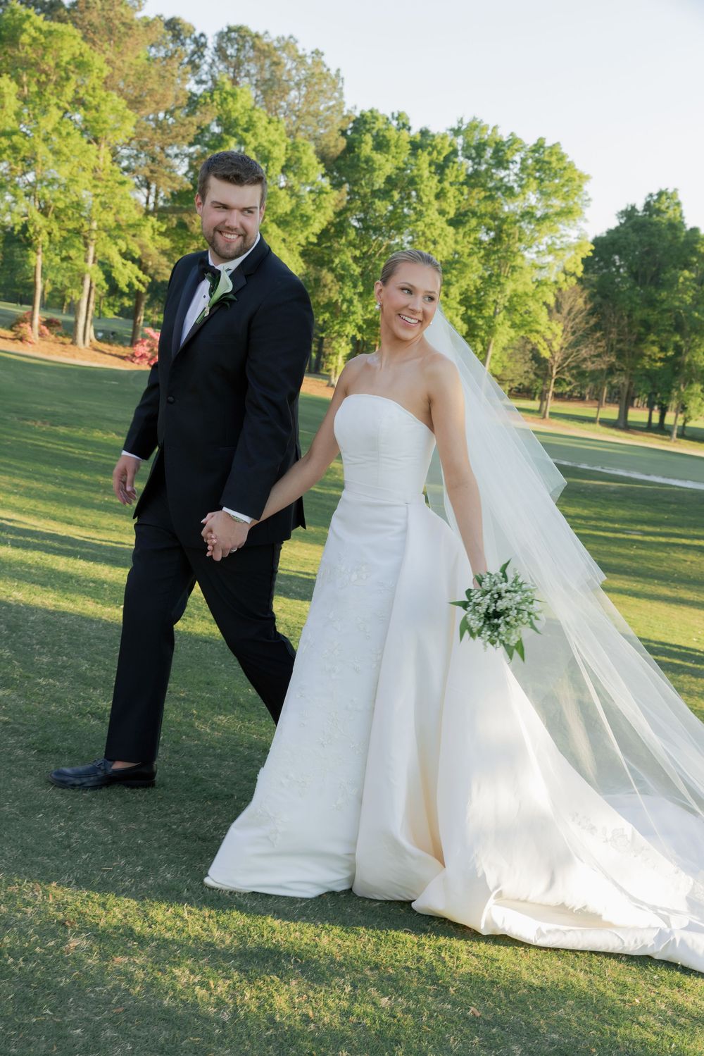 Bride and groom walk hand-in-hand across a lush green golf course on their wedding day.