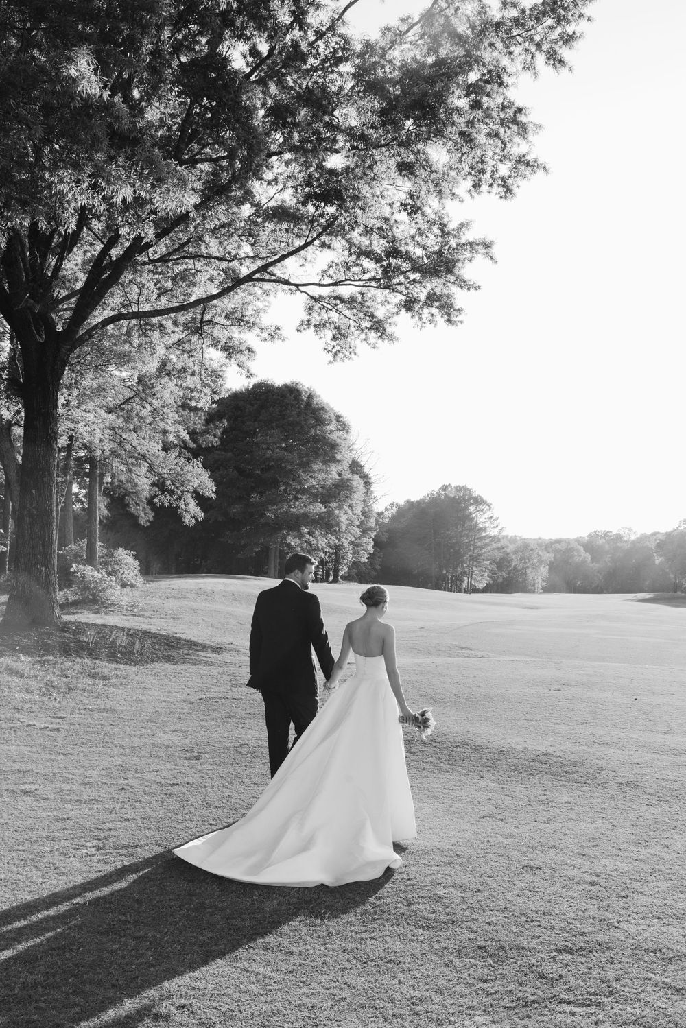 Bride and groom walk hand-in-hand across sunlit golf course in romantic black and white wedding portrait.