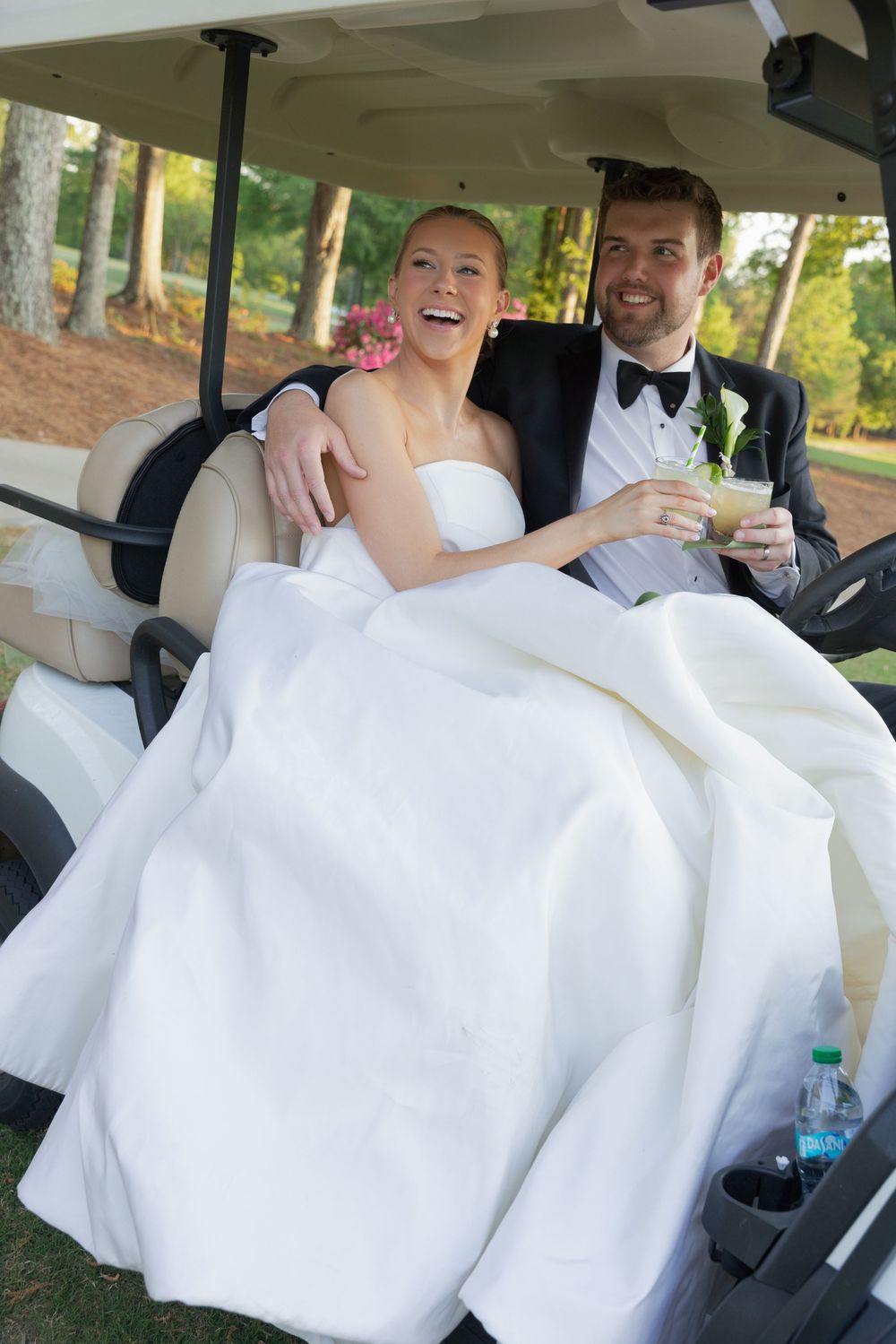 Happy bride and groom riding in a golf cart on their wedding day, smiling and holding drinks outdoors.