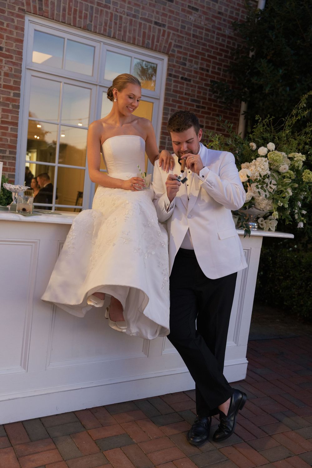 Bride sitting on outdoor bar ledge while groom kisses her hand at elegant wedding reception.