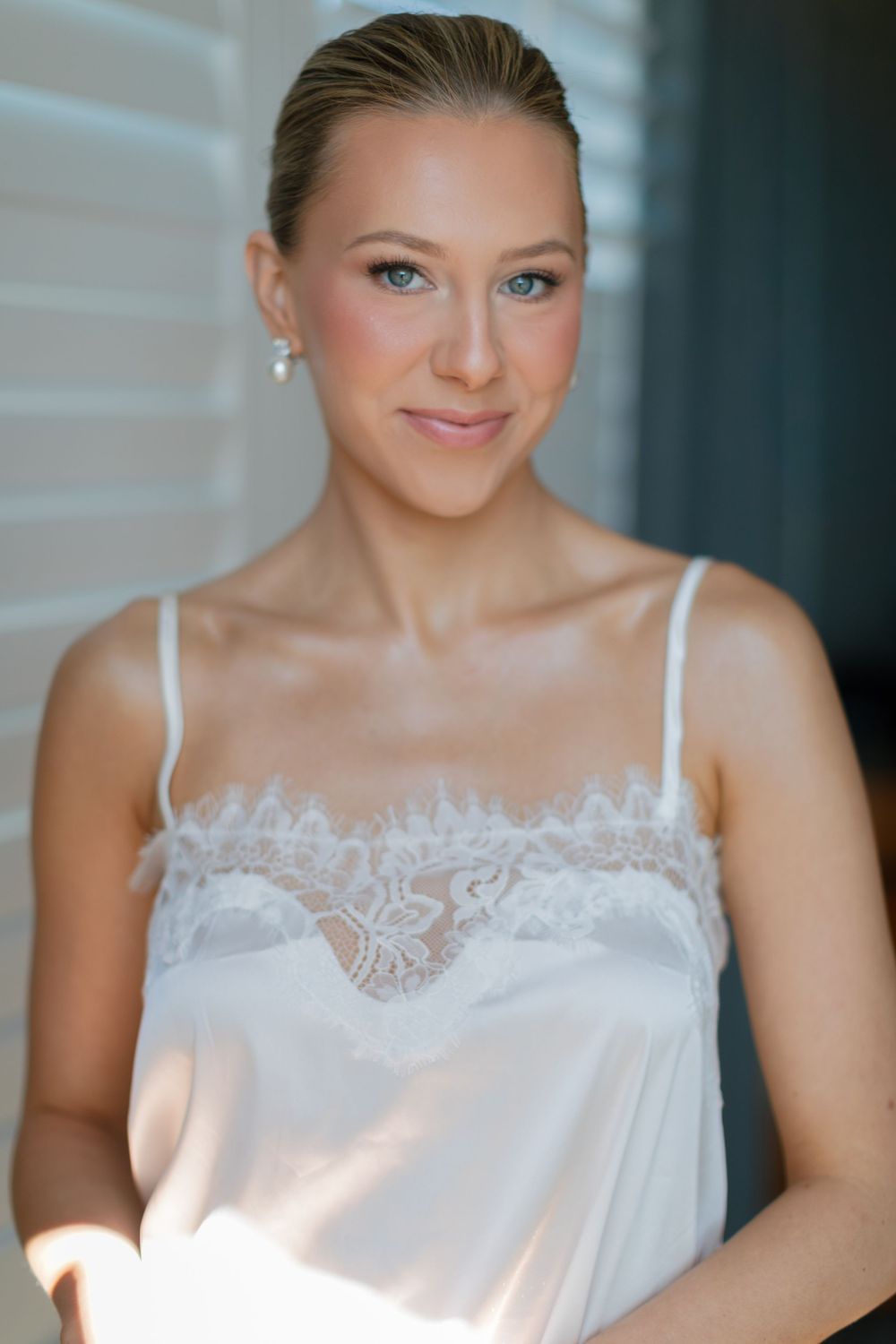A young woman in a white lace camisole with pearl earrings smiles softly, with natural makeup and sleek hair.