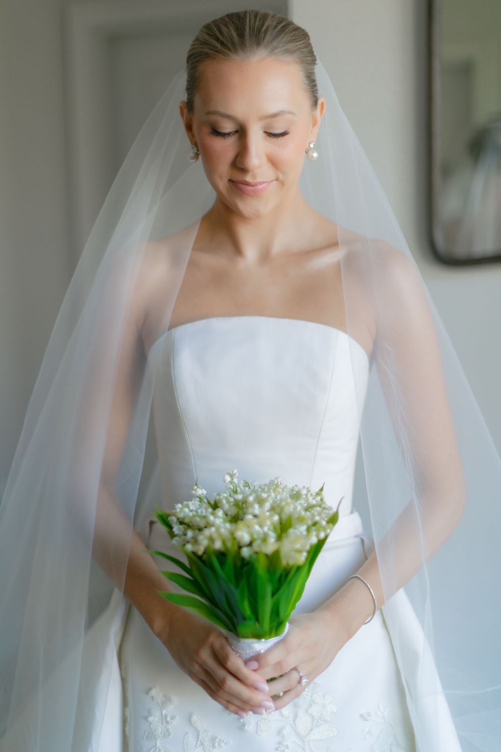 Bride in strapless white gown and veil holds lily of the valley bouquet with eyes closed.
