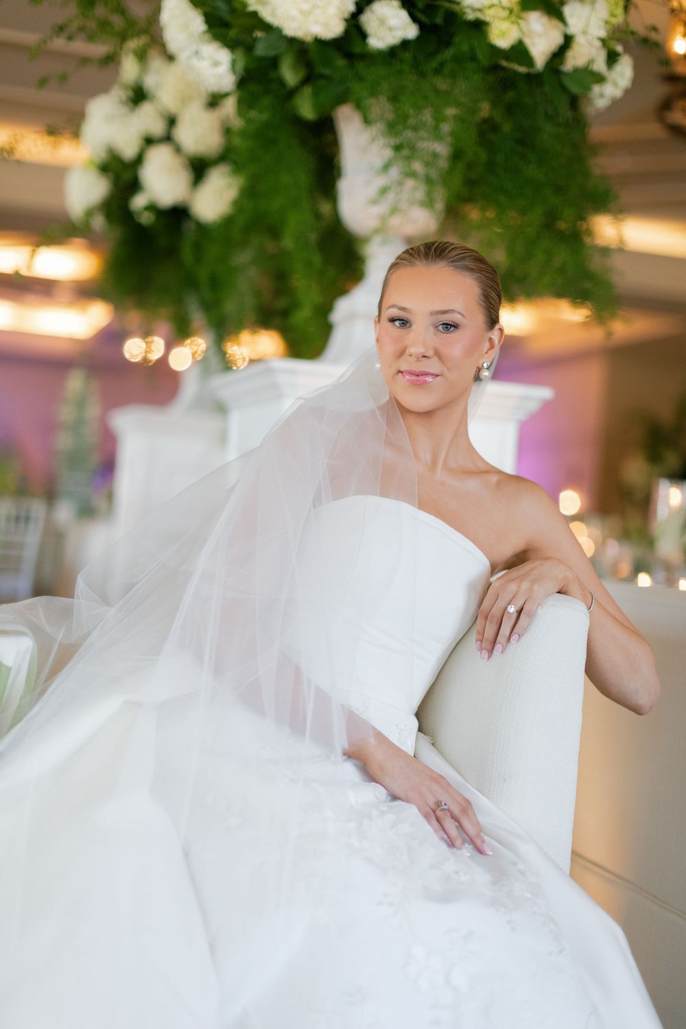 Elegant bride in a strapless white wedding gown and veil sits gracefully in a white chair at a floral reception venue.