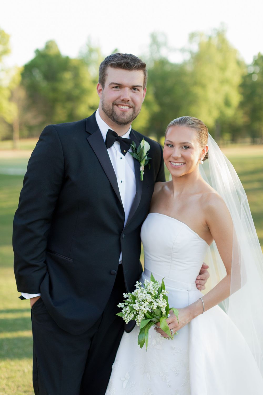 Smiling bride and groom pose outdoors on golf course, bride holding lily of the valley bouquet.