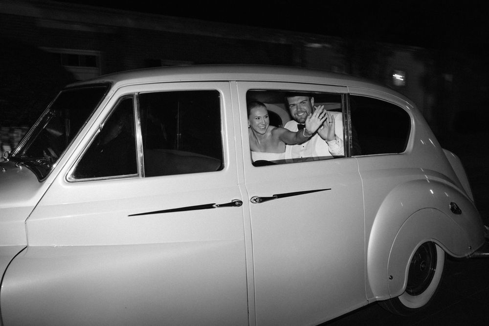 Black and white photo of a bride and groom inside a vintage white car at night.