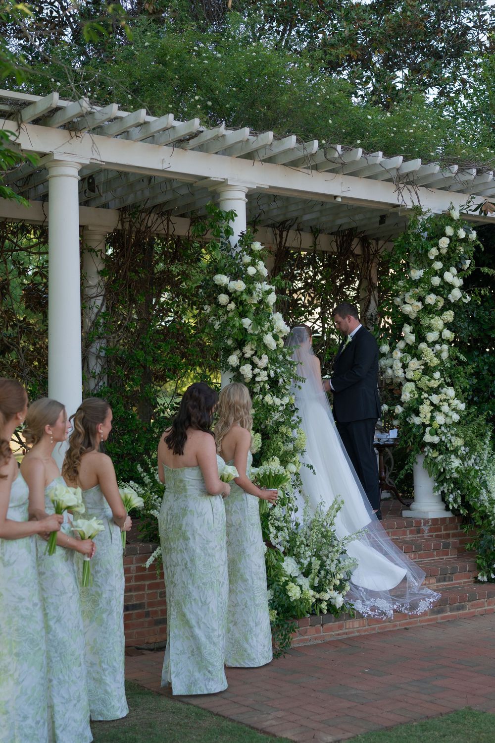Bride and groom exchange vows under white pergola adorned with white floral arrangements as bridesmaids look on.