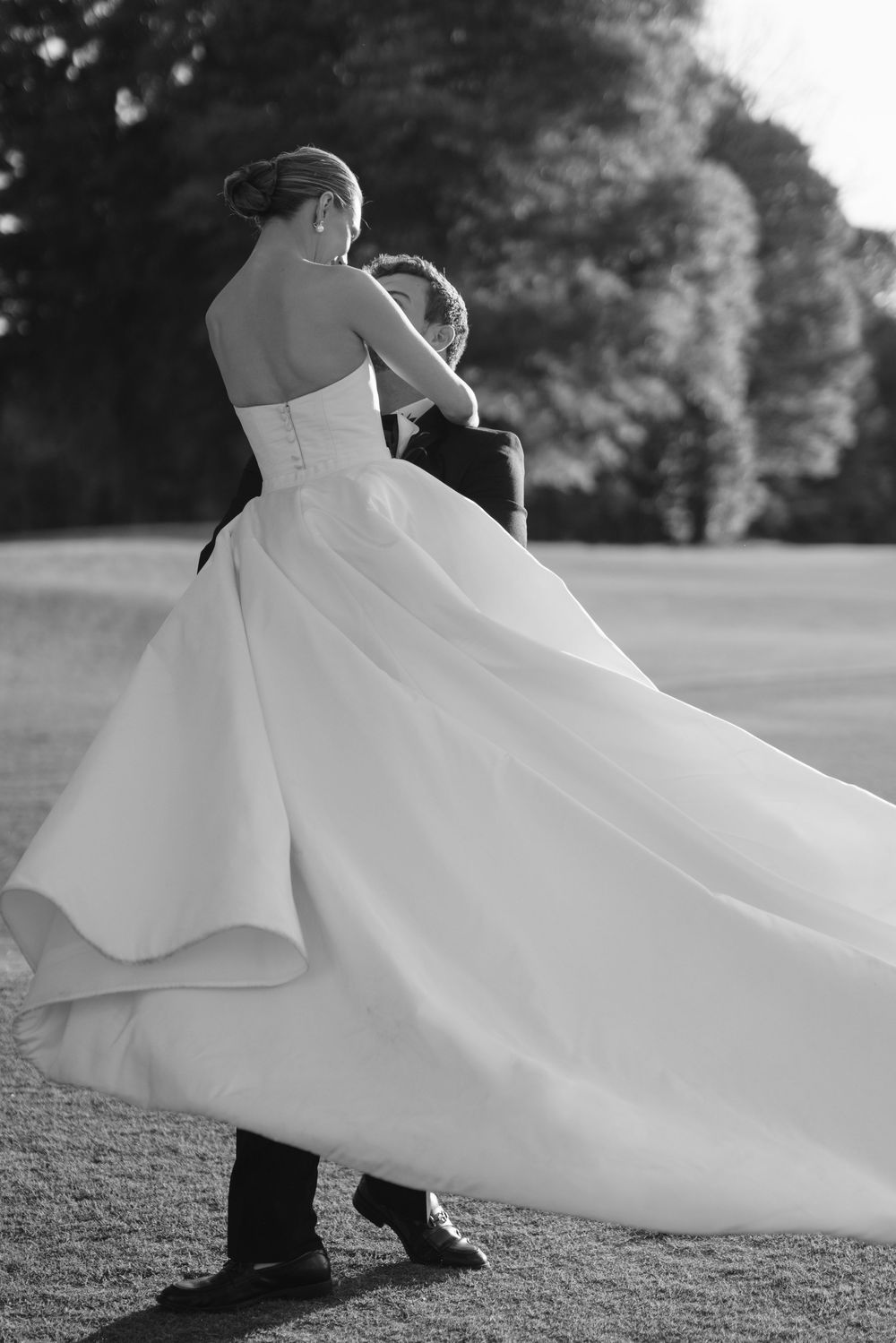 Black and white photo of bride in flowing strapless ball gown dancing with groom outdoors on grass.