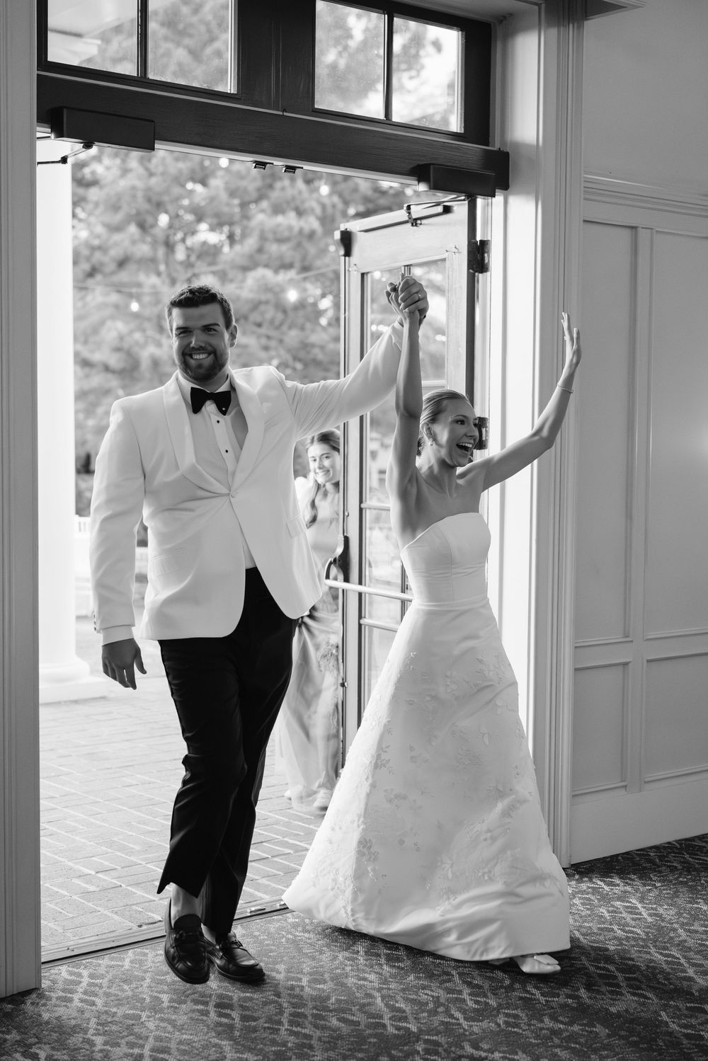Black and white photo of smiling bride and groom making grand entrance through venue doors hands raised.
