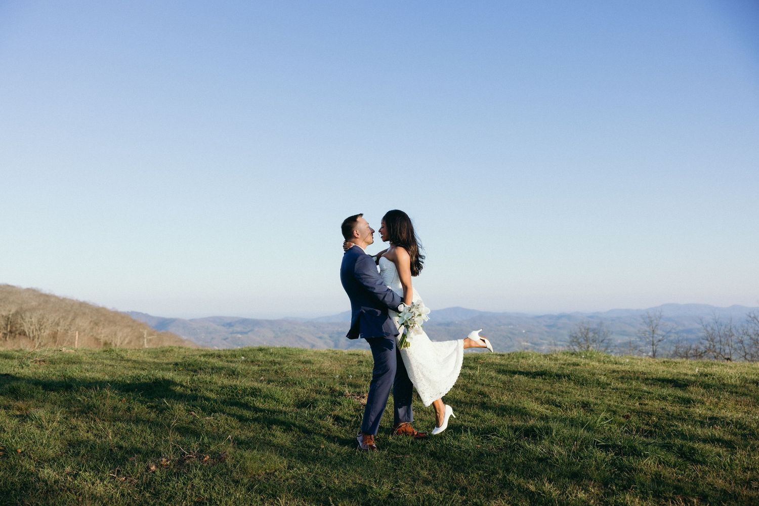 Couple embracing on a scenic hilltop, groom lifting bride in white dress with mountain views under clear blue sky.