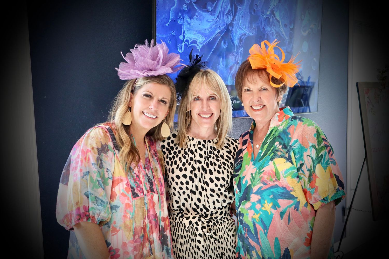 Three beautiful smiling women wearing colorful floral outfits and decorative hats pose together at an event.