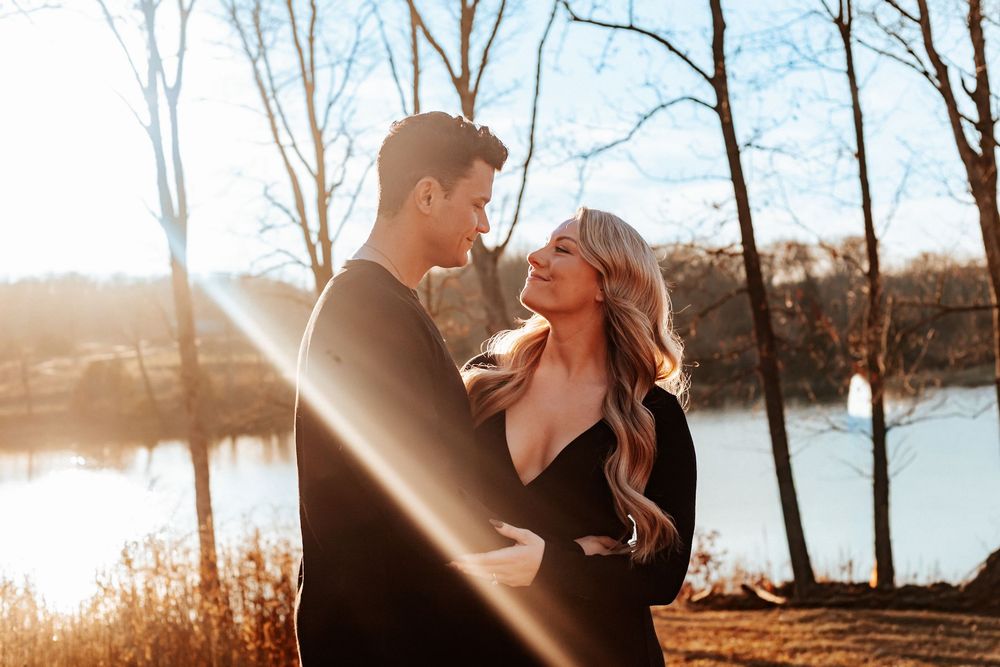 Couple in black outfits embracing near a sunlit lake with bare trees in the background.