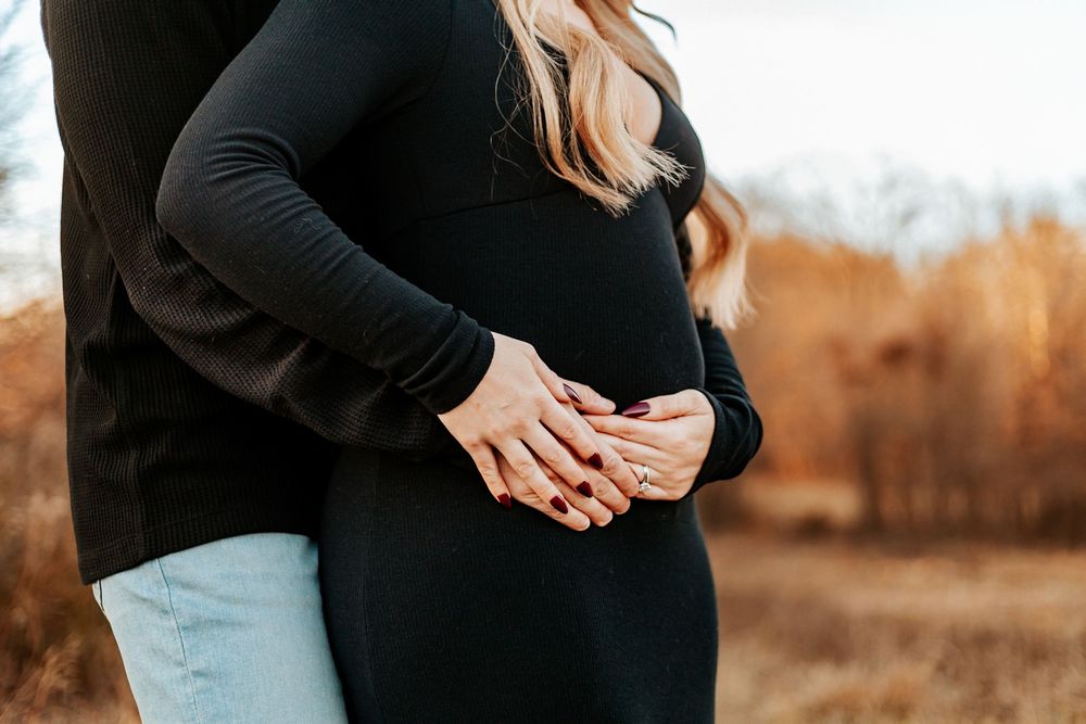 Pregnant woman in black dress with partner's hands gently cradling her baby bump outdoors.