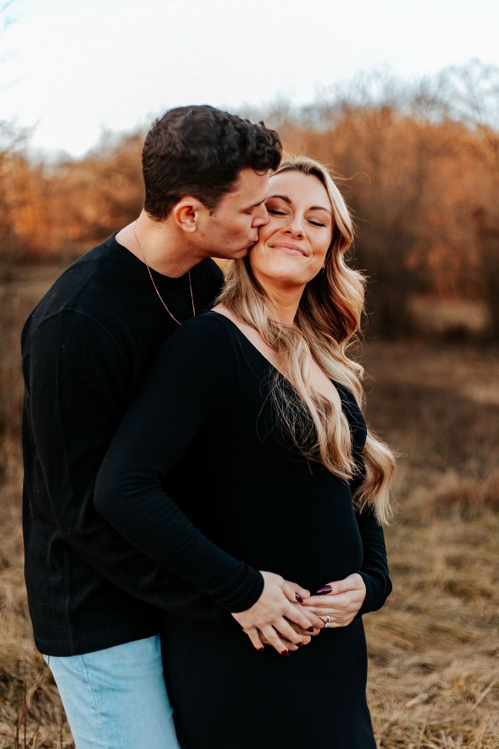 Couple in black outfits sharing a tender moment in autumn field, man kissing pregnant woman's cheek.