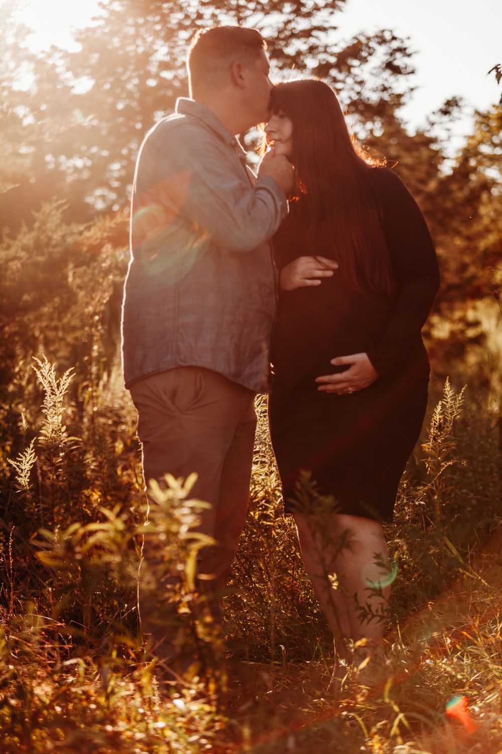 Couple sharing a kiss in golden sunset light, woman in black dress showing baby bump.