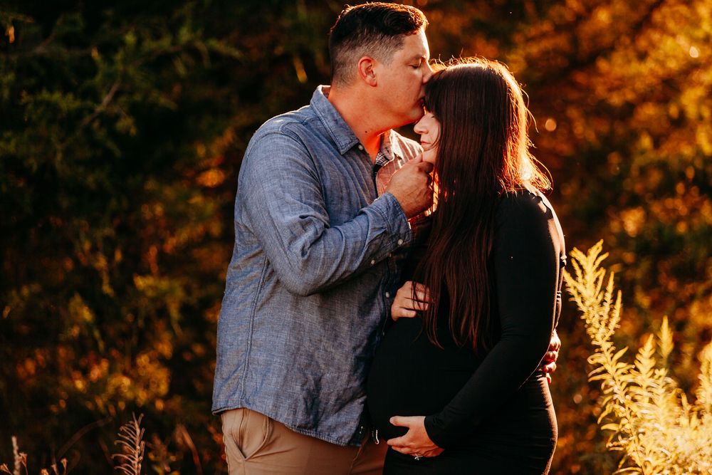Couple sharing a tender kiss during golden hour maternity photoshoot outdoors with autumn foliage background.