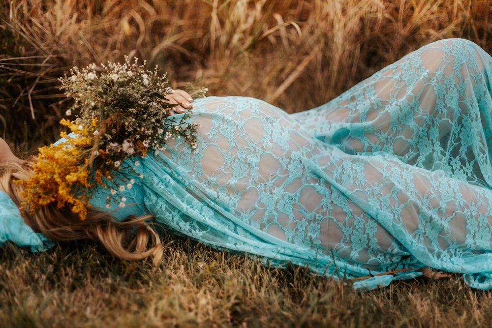 Woman in teal lace gown lying in golden field holding wildflower bouquet with floral crown in hair.