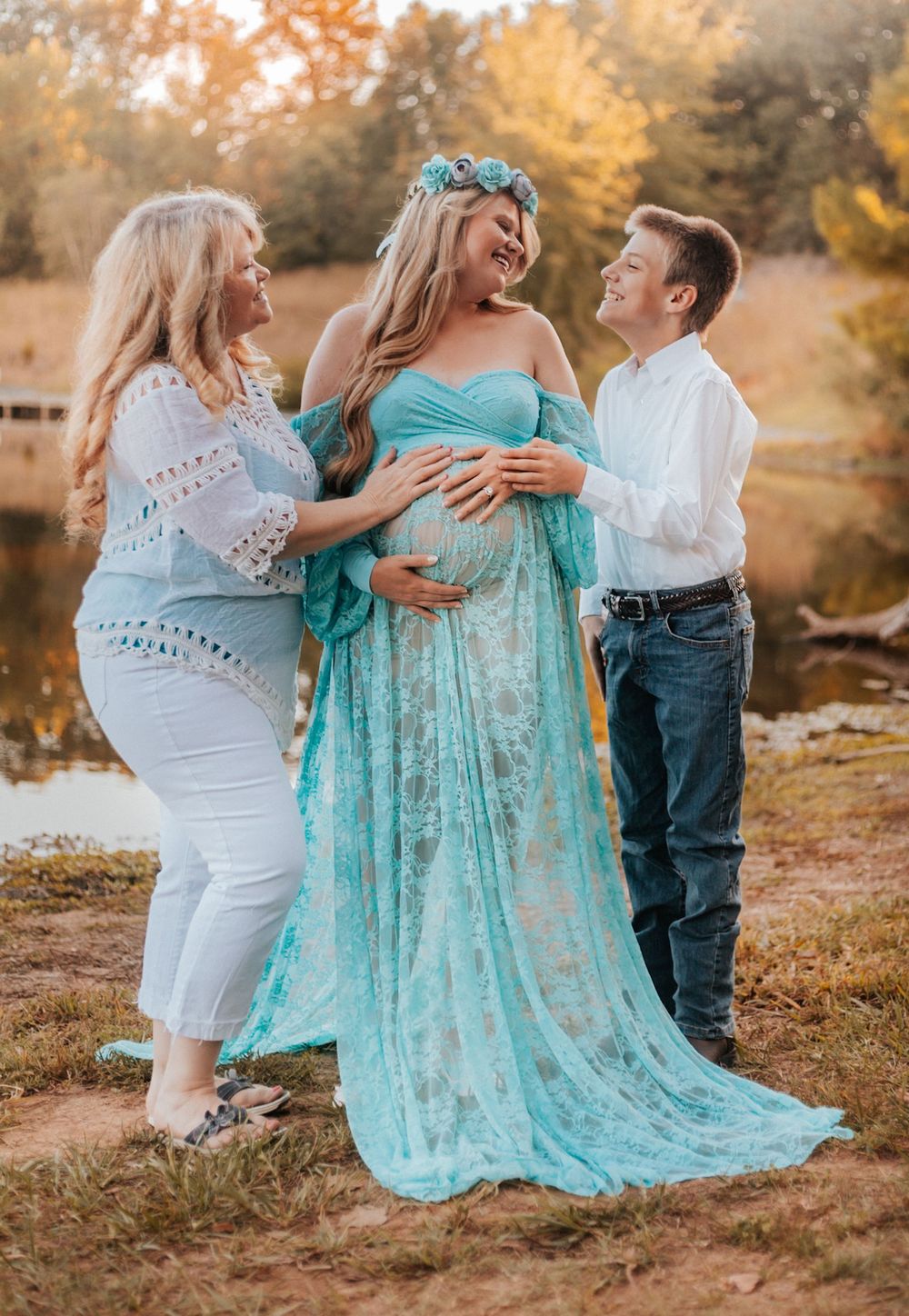 Pregnant woman in teal lace gown with floral crown poses outdoors with two smiling companions during maternity photo session.